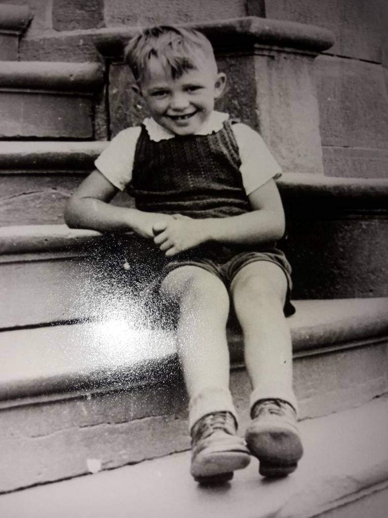Schwarz/Weiß-Foto. Ein kleiner Junge mit einer kurzen Latzhose sitzt auf einer Steintreppe.
